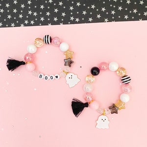 May include: Two pink and black Halloween themed bracelets with ghost charms and "BOO" beads. The bracelets have black tassels and are on a pink background.