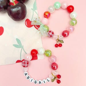 May include: Two beaded bracelets with the name "ABIGAIL" spelled out in white letter beads. The bracelets feature a variety of colorful beads, including red, pink, green, and iridescent beads. Each bracelet has a cherry charm and a green bow. Fresh cherries are in the background.