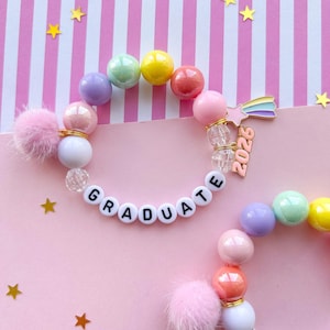 May include: Two colorful beaded bracelets with the word "GRADUATE" spelled out in white letter beads. The bracelets feature pastel-colored beads, fluffy pink pom-poms, and a rainbow star charm with "2026" on it. Gold star confetti.