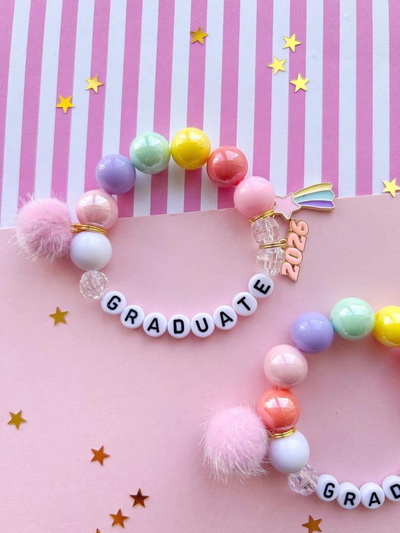 May include: Two colorful beaded bracelets with the word "GRADUATE" spelled out in white letter beads. The bracelets feature pastel-colored beads, fluffy pink pom-poms, and a rainbow star charm with "2026" on it. Gold star confetti.