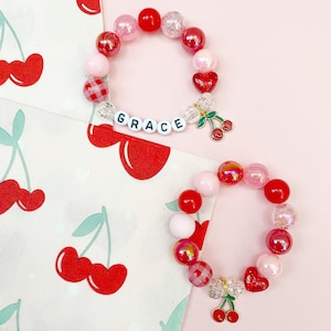 May include: Two red and pink beaded bracelets with a cherry charm. The top bracelet has the name "GRACE" spelled out in white letters. The bracelets are on a pink background with a white background with red cherries.