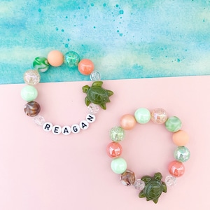 May include: Two colorful beaded bracelets with a turtle charm. The top bracelet has the name "REAGAN" spelled out in white beads. The bracelets are on a pink background.