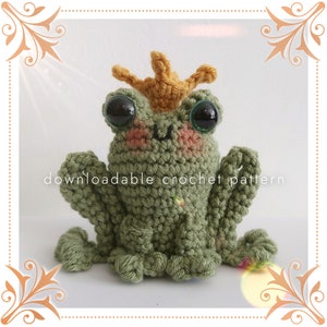 May include: A green crocheted frog wearing a yellow crocheted crown. The frog has large black eyes and a pink blush on its cheeks. The frog is sitting on a white background. "Downloadable crochet pattern" is written in black text below the frog.