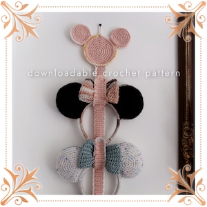 Puede incluir: Un soporte para diademas de crochet rosa con una cabeza de Mickey Mouse de crochet rosa y amarilla en la parte superior. El soporte tiene tres diademas de crochet con lazos colgando. Las diademas son negras, marrón y blancas, y azul verdoso y blancas.