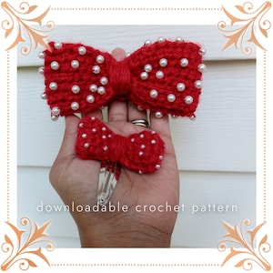 May include: Two red crocheted hair bows with white pearl embellishments. The larger bow is shown on a hand, while the smaller bow is attached to a silver alligator clip.