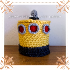 May include: A yellow crocheted toilet paper roll cover with a gray and red trim and a small gray pom-pom on top. The text "downloadable crochet pattern" is visible at the top of the image.