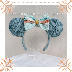 Può includere: Fascia per capelli a forma di orecchie di topo in crochet di colore azzurro chiaro con un fiocco multicolore. Il fiocco è realizzato in filato azzurro chiaro, bianco e arancione con un accento dorato. La fascia è su un supporto trasparente.