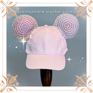 May include: A pink baseball cap with two crocheted rainbow ears. The ears are attached to the top of the cap. The cap has a white brim and a white crown. The cap is on a wooden stand.
