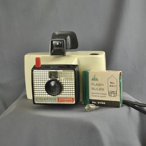 May include: A vintage Polaroid Swinger Model 32 camera with a flash bulb and a box of flash bulbs. The camera is white with a black lens and a red button. The box of flash bulbs is green and white with the text "Flash Bulbs No. AG-1 pocket-sized twelve-pack No. 8782".
