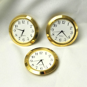 May include: Three gold-tone quartz clock inserts with white faces and black hands. The clock faces have black numerals and the word "QUARTZ" printed at the bottom. The clocks are circular and have a gold-colored bezel.