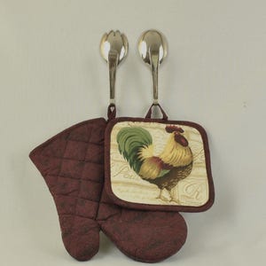 May include: A set of two red quilted oven mitts with a rooster design pot holder. The pot holder features a rooster with a green, yellow, and brown tail and a white background with French text. The text reads "J'ai peur de la chaleur" which translates to "I am afraid of the heat".