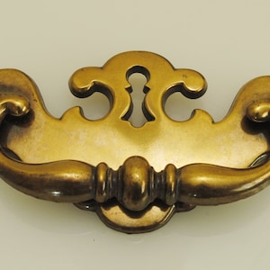 May include: A brass cabinet pull with a decorative, ornate design. The pull has a keyhole in the center and a curved handle.