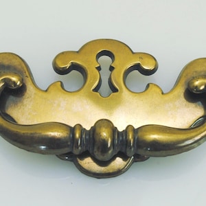 May include: A brass drawer pull featuring a curved, swinging handle attached to a decorative backplate. The backplate has a central keyhole cutout and symmetrical, scrolling edges. The metal has a polished, aged finish with visible highlights.