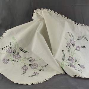 May include: A white crocheted tablecloth with a scalloped edge and embroidered purple flowers with black stems and leaves.