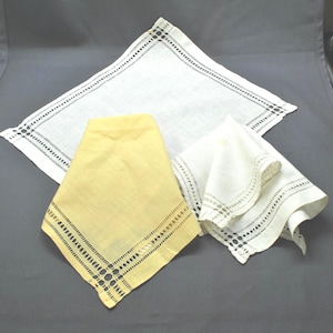 May include: Four vintage handkerchiefs in the image. One is yellow, and the others are white. Each handkerchief has a decorative border with a cut-out design. The handkerchiefs are made of a soft, likely linen material.