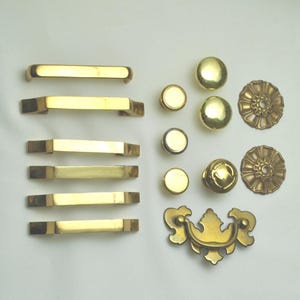 May include: A collection of brass hardware, including cabinet pulls, knobs, and decorative rosettes. The pulls are rectangular and bar-shaped, while the knobs are round. The rosettes have a floral design. All pieces have a shiny, golden finish.