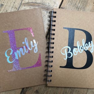 May include: Two brown spiral notebooks with personalised names. The notebook on the left has a pink glitter letter "E" with the name "Emily" in white script. The notebook on the right has a black letter "B" with the name "Bobby" in white script.