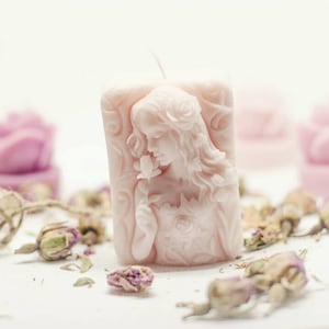 Op de afbeelding: Een roze kaars met een gedetailleerd beeldhouwwerk van een vrouw die een bloem vasthoudt. De kaars is omringd door gedroogde rozenknoppen.