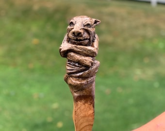 May include: A wooden walking stick with a carved animal head at the top. The head is brown and has a detailed face with a mischievous expression. The stick is made of light brown wood and has a smooth finish.