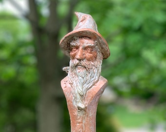 May include: A terracotta sculpture of a wizard with a long white beard and a pointed hat. The sculpture is weathered and has a rustic appearance.