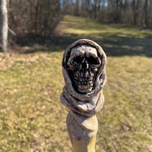 May include: A detailed, hand-carved wooden walking stick with a skull head at the top. The skull is wearing a hooded robe and has a weathered, aged look.