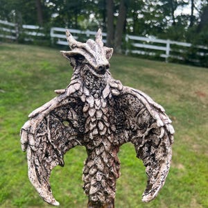 May include: A detailed sculpture of a dragon with outstretched wings. The dragon is primarily off-white with dark brown accents, and the wings have a textured, feathered appearance. The sculpture is mounted on a wooden staff, creating a unique fantasy art piece.