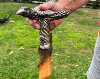 May include: A wooden walking cane with a carved bird-shaped handle. The handle is a dark, textured silver color, transitioning to a twisted, silver and brown shaft. The lower portion of the cane is a smooth, light brown wood.