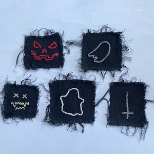 May include: Five black fabric patches with frayed edges, each featuring embroidered designs. The designs include a jack-o'-lantern face, a ghost, a wing, a ghost, and an upside-down cross. The embroidery colours are red, white, and beige.