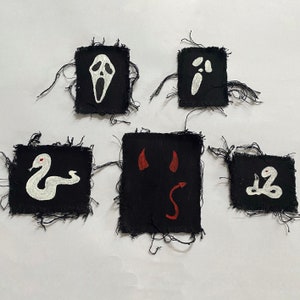 Peut inclure: Cinq patchs en tissu noir avec des motifs brodés blancs et rouges. Les motifs comprennent un visage de fantôme, un visage de diable, un serpent, un crâne et un numéro 2.