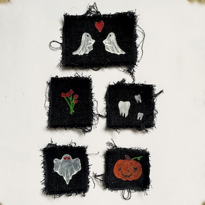 May include: Five small, black fabric squares with embroidered designs. The designs include two white ghosts with a red heart, red flowers, white teeth, a ghost with red eyes, and a jack-o'-lantern. The edges of the squares are frayed.