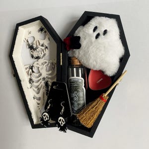 Könnte beinhalten: Eine offene, schwarze Sarg-Box mit Halloween-Themen. Im Inneren befinden sich ein Skelett, ein Plüschgeist, eine Flasche mit Etikett, eine herzförmige rote Kerze, ein kleiner Besen und Geisterohrringe.