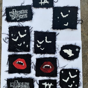 May include: Nine black fabric patches with white embroidered designs. The patches feature the words "Motionless in White", a Roman numeral "IX", and bats. Two patches feature a red, open mouth with fangs.