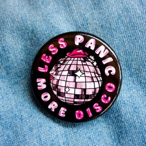 May include: A round, black button with the text "LESS PANIC MORE DISCO" in pink lettering. The button features a pink and white disco ball wearing a pink hat. The button is set against a blue denim background.