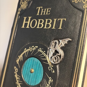 Op de afbeelding: Een zwart leren boek met de titel "The Hobbit" met gouden letters. De kaft heeft een turquoise deur, een zilveren draakspeld en gouden cirkelvormige tekst. Het boek heeft gouden randen en hoekontwerpen.