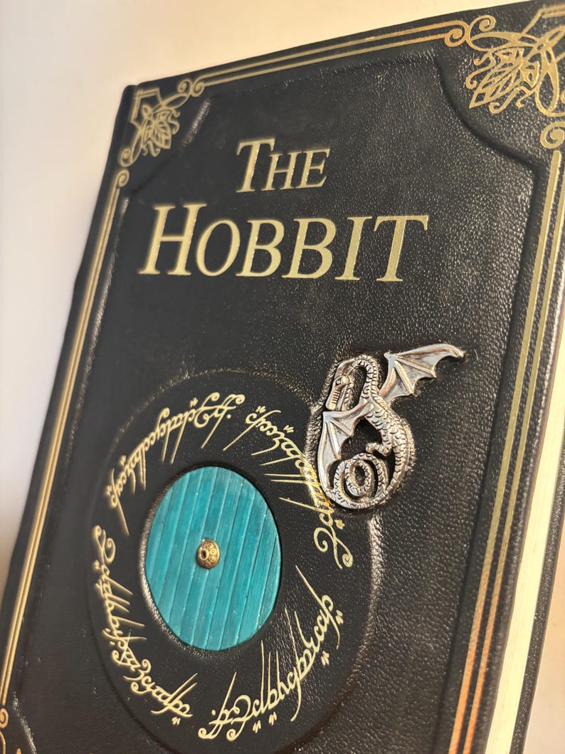 Op de afbeelding: Een zwart leren boek met de titel "The Hobbit" met gouden letters. De kaft heeft een turquoise deur, een zilveren draakspeld en gouden cirkelvormige tekst. Het boek heeft gouden randen en hoekontwerpen.