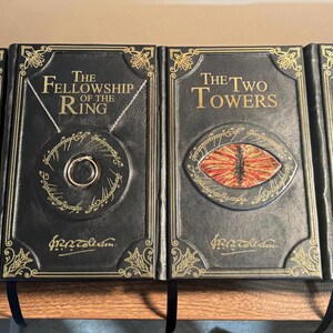 Op de afbeelding: Vier hardcover boeken uit de Lord of the Rings-serie. De boeken zijn zwart met gouden letters en decoratieve accenten. De titels zijn "The Hobbit", "The Fellowship of the Ring", "The Two Towers" en "The Return of the King".