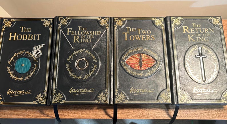 Op de afbeelding: Vier hardcover boeken uit de Lord of the Rings-serie. De boeken zijn zwart met gouden letters en decoratieve accenten. De titels zijn "The Hobbit", "The Fellowship of the Ring", "The Two Towers" en "The Return of the King".