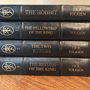 Op de afbeelding: Een stapel van vier zwarte hardcover boeken van J.R.R. Tolkien. De titels zijn "The Hobbit", "The Fellowship of the Ring", "The Two Towers" en "The Return of the King". Gouden letters en een decoratief embleem op de ruggen.