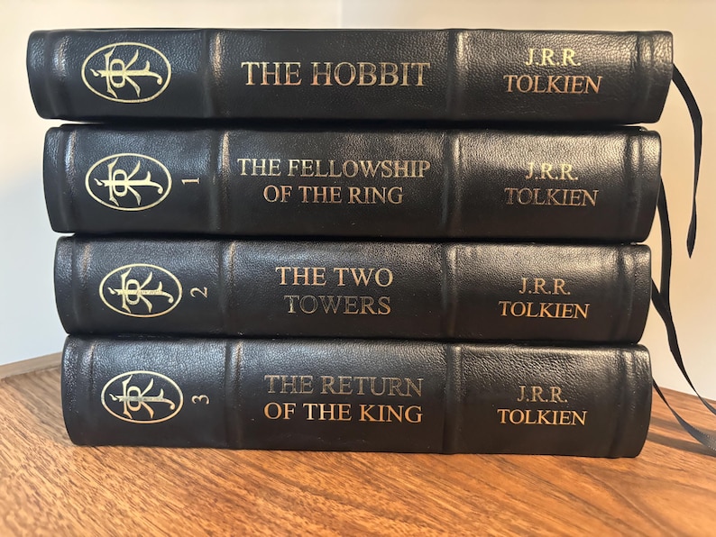 Op de afbeelding: Een stapel van vier zwarte hardcover boeken van J.R.R. Tolkien. De titels zijn "The Hobbit", "The Fellowship of the Ring", "The Two Towers" en "The Return of the King". Gouden letters en een decoratief embleem op de ruggen.