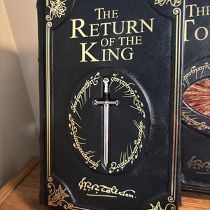 Op de afbeelding: Een zwart boek getiteld "The Return of the King" met gouden letters en accenten. Een zilveren zwaard is gecentreerd in een gouden ringontwerp. Het boek heeft decoratieve hoekontwerpen.