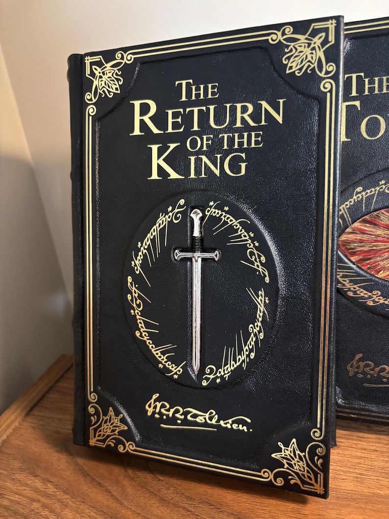 Op de afbeelding: Een zwart boek getiteld "The Return of the King" met gouden letters en accenten. Een zilveren zwaard is gecentreerd in een gouden ringontwerp. Het boek heeft decoratieve hoekontwerpen.