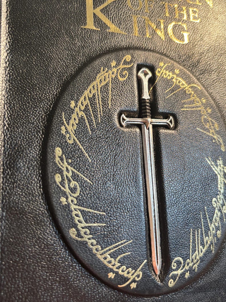 Op de afbeelding: Close-up van een zwarte leren boekomslag met gouden letters met de tekst "The King". Een zilveren zwaard is gecentreerd op een cirkelvormig embleem met gouden letters. De boekomslag heeft een getextureerde uitstraling.