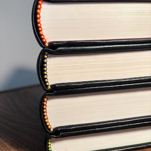 Op de afbeelding: Een stapel van drie boeken met zwarte leren covers. De randen van de pagina's zijn wit en de ruggen zijn versierd met kleurrijke stiksels in rood, geel, blauw en groen. De boeken liggen op een houten oppervlak.