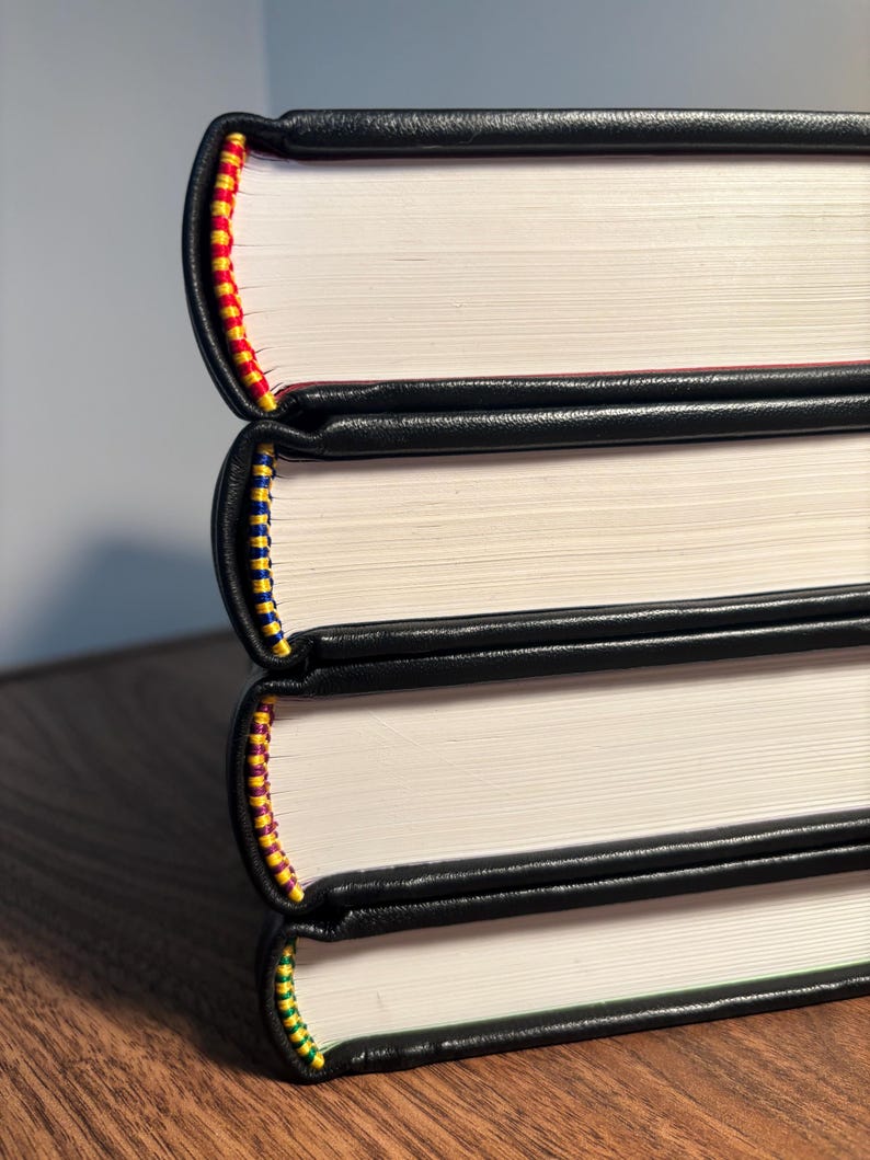 Op de afbeelding: Een stapel van drie boeken met zwarte leren covers. De randen van de pagina's zijn wit en de ruggen zijn versierd met kleurrijke stiksels in rood, geel, blauw en groen. De boeken liggen op een houten oppervlak.