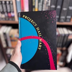 May include: A hardcover book titled Project Hail Mary with a black cover featuring a large blue circle, a curved magenta stripe, and small pink star-like accents. The title is printed in yellow capital letters along the curved blue edge.