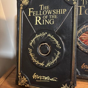 Op de afbeelding: Een zwart leren boek met de titel "The Fellowship of the Ring" met gouden letters en details. Een zilveren ketting houdt een cirkelvormige hanger met een gouden ring vast. De hanger heeft inscripties rondom.
