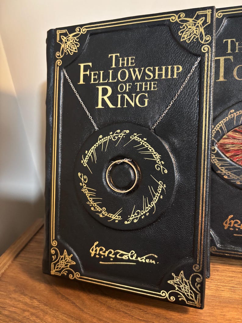 Op de afbeelding: Een zwart leren boek met de titel "The Fellowship of the Ring" met gouden letters en details. Een zilveren ketting houdt een cirkelvormige hanger met een gouden ring vast. De hanger heeft inscripties rondom.