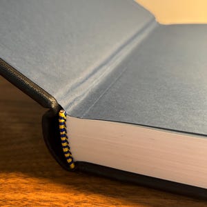 Op de afbeelding: Een open boek met een blauwe kaft en een zwarte rug. De pagina's zijn wit en de binding is een geel en blauw gestreept lint. Het boek ligt op een houten oppervlak.
