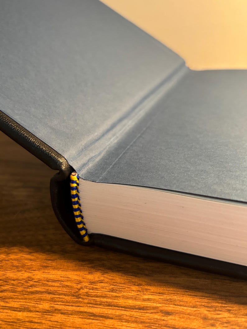 Op de afbeelding: Een open boek met een blauwe kaft en een zwarte rug. De pagina's zijn wit en de binding is een geel en blauw gestreept lint. Het boek ligt op een houten oppervlak.