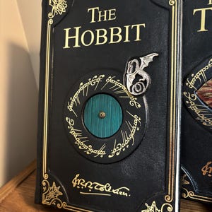 Op de afbeelding: Een zwart leren boek met de titel "The Hobbit" met gouden letters en accenten. De omslag heeft een cirkelvormig ontwerp met een groene deur, een zilveren draak en inscripties. Het boek wordt gepresenteerd op een houten oppervlak.